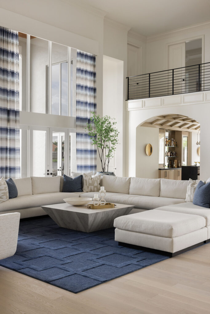 High-ceiling living room featuring a large cream sectional sofa arranged on a textured navy blue rug with a faceted coffee table at its center. Tall blue-and-white striped drapes frame floor-to-ceiling windows, while an archway in the background leads to a bar area below a second-floor loft.