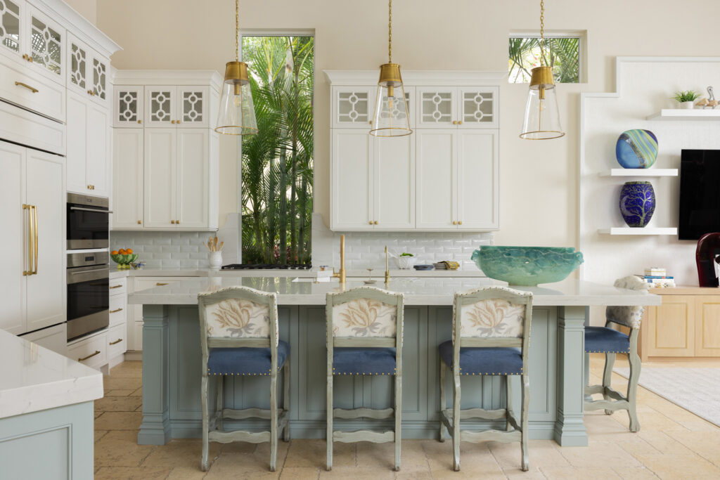 Bright, open-concept coastal kitchen with a large blue island, white cabinets with gold hardware, and a tall window overlooking tropical foliage