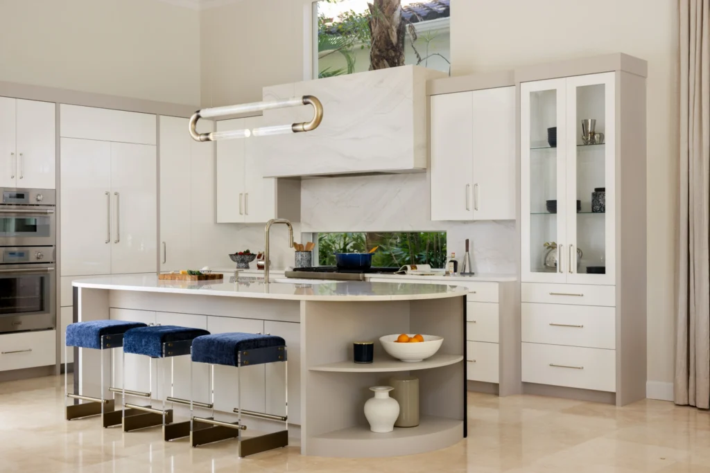 Modern kitchen with off-white cabinetry, a marble range hood, and a kitchen island featuring a curved open-shelf end. Three blue velvet barstools with clear acrylic legs tuck under the island beneath a modern tubular brass pendant light, flanked by a tall glass-front display cabinet.