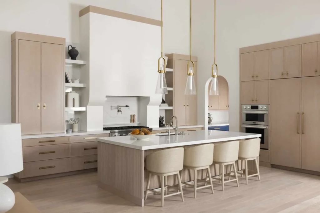 Light, neutral kitchen with custom beige cabinetry, large white island with seating, brass pendant lights, and integrated built-in appliances in a modern luxury interior.